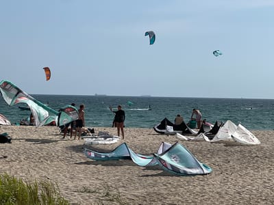 The image depicts a crowded beach scene, likely in Tarifa, Spain, renowned for its kiteboarding conditions. The foreground is dominated by a sandy beach, with several large kites spread out on the sand, indicating preparations for or aftermath of kiteboarding activities. Some kites are partially inflated, while others are lying flat. In the middle ground, a group of people are present on the beach, some standing and observing, others possibly involved with the kites. Two individuals in the center are near a large, partially inflated kite with the number "10" visible. Further back, the turquoise water of the sea is visible, with several kiteboarders actively engaged in the sport in the distance. The sky is clear and blue, with a few distant ships on the horizon. The overall atmosphere suggests a busy day for kiteboarding enthusiasts in a popular coastal location.