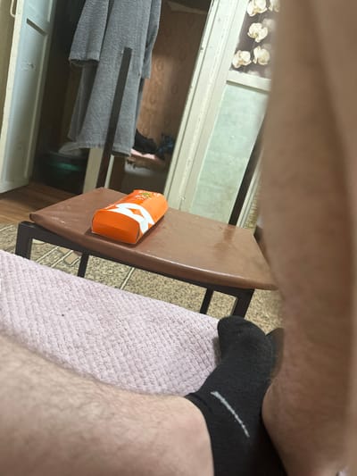 The image shows a close-up of a person's legs and feet. The person is wearing black socks. In the background, there is a small brown table with an orange box on it. Behind the table, there is a closet with clothes hanging inside. The location is in the city of Corlăteni, Moldova.