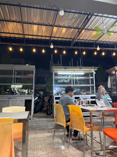 Here is a description of the image:

The image depicts an outdoor evening scene, likely in a roadside food stall in Kutalhoksukon, Indonesia. The focus is on a seating area with several orange and yellow plastic chairs positioned around metal tables. A man wearing a grey t-shirt is seated at one of the tables, looking at a phone. Another man lies down on the same table, apparently resting.

The stall is illuminated by strings of lights and an electric fan is visible under the corrugated metal roof. There are other food stalls and parked motorcycles in the background.