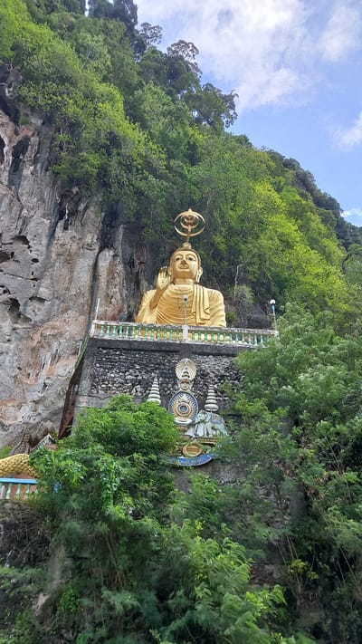 A statue of Buddha rests upon a rugged, grey rock face in Thap Put, Thailand. The statue is gold, with a halo-like structure above its head. It stands behind a short, greenish-blue decorative wall atop a pile of jagged rocks. Lush green vegetation surrounds the statue, covering the upper portion of the rock face and obscuring much of the backdrop. The sky is partly visible in the upper left, a pale blue with some white clouds.