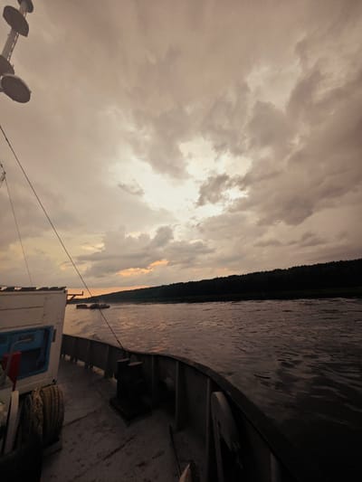 This media file depicts a view from the deck of a boat sailing on a river, likely in Kazachinskoe, Russia. The time of day appears to be either dawn or dusk, as suggested by the warm, muted tones of the sky and the soft lighting. The sky is overcast with dramatic clouds, creating a somber or contemplative mood.
The foreground of the image shows a portion of the boat's deck, with a coil of thick rope prominently displayed to the left and a dark bollard situated in the center. A white structure with a blue opening is also visible on the deck. A dark rope stretches diagonally across the frame, connecting to unseen parts of the boat.
In the mid-ground, the river surface is textured with ripples, reflecting the muted light. Further in the distance, another boat, possibly a tugboat, is visible. The far bank of the river is lined with a dense, dark forest, creating a stark silhouette against the sky.
The overall impression is one of solitude and the quiet movement of a vessel through a natural landscape under a dramatic sky. There is no visible text in the image.