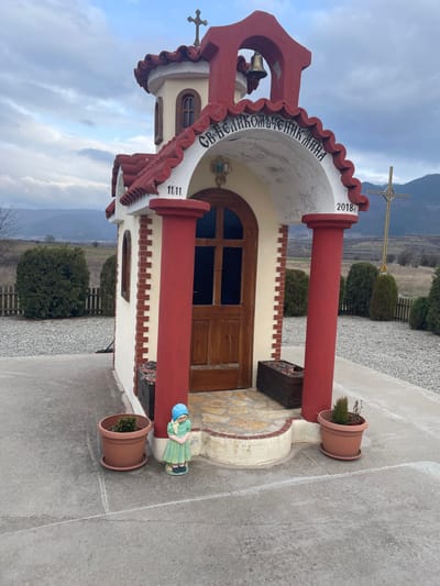 The scene captures a small, cream-colored, single-story chapel-like structure situated on a concrete pad in Krainitsi, Bulgaria. The building features a prominent red-tiled roof with a small belfry containing a gold-colored bell and topped by a cross. Its facade includes an arched entryway supported by two prominent red pillars, framing a dark wooden door with four vertical glass panels. Above the door, Cyrillic text reads "СВ. ВЕЛИКОМЪЧЕНИК МИНА", flanked by "11.11." on the left and "2018г." on the right. A small icon is centered above the door within the arch. To the left of the entrance, a small, light-green female figurine with a blue hat stands next to a terracotta-colored potted plant containing a small green shrub. Another similar potted plant is to the right of the entrance. A dark wooden planter box sits to the right of the doorway. The structure's sides feature red brick-like detailing. The immediate ground around the concrete pad is covered in gravel, bordered by a low wooden fence. Beyond the fence, green shrubs and open, uncultivated fields extend towards distant mountains. A tall, golden, plain cross on a stand is visible to the right of the chapel in the mid-ground. The sky is overcast with varying shades of gray and blue.
