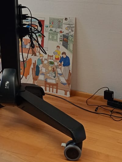 A still, indoor photograph captures a scene on a wooden floor. In the foreground, a black, wheeled stand, likely for a television or monitor, is angled towards the right. Cables are visible trailing from the stand.
To the right of the stand, a colorful, framed picture or canvas is propped against a light-colored wall. The artwork depicts a cartoonish scene of a family gathered around a dining table in a kitchen. Several people are illustrated, some setting the table, while others are seated. A turkey is visible in an oven in the background.
Further to the right, against the same wall, a black power strip with several outlets is placed on the floor. A black power adapter is plugged into one of the outlets, with its cable extending towards the left. Other black cables are also visible on the floor, connecting to the power strip and potentially to the stand.
The overall impression is a domestic setting, possibly a living room or den, with a focus on technology and a piece of artwork. There are no obvious cues for time of day or weather. The mood is neutral. No visible text is discernable in the image, other than what appears to be a small logo on the stand.