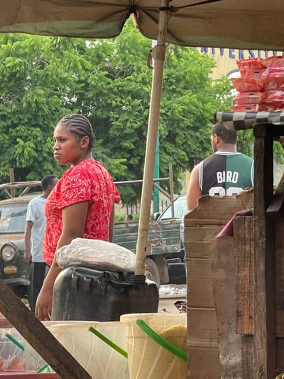 The image depicts an outdoor market or roadside stall during the daytime, characterized by a rustic setup and several individuals. The primary orientation of the image is rotated 90 degrees counter-clockwise; for this description, it will be analyzed as if correctly oriented.
In the foreground, on the left, an olive-green canvas awning provides shade, supported by visible poles. To the right of the awning, a young woman stands behind what appears to be a stall counter, made of rough wood and possibly cardboard. She is wearing a red short-sleeved top with a white patterned design and has her dark hair styled in intricate braids. Her gaze is directed downwards, suggesting she is focused on an item or task in front of her.
Immediately to her right, on the stall counter, sits a large, black plastic container, possibly a jerry can or a drum, with a white plastic bag covering part of its opening. Further to the right, a light-colored plastic bucket with a green handle and spout is visible, containing a granular, pale yellow substance, likely grains such as rice or maize.
In the midground, behind the stall, another person, a man, is partially visible. He is wearing a green, black, and white basketball jersey with "BIRD" and the number "33" clearly visible on the back. His back is mostly turned towards the viewer, and he has short, dark hair or a beard. He is standing behind a cardboard barrier, possibly another part of a stall or a display.
The background is dominated by lush, green foliage from numerous trees, indicating an outdoor, possibly natural, setting. Behind the woman and the black container, a portion of an older, dark green utility vehicle, possibly a truck or jeep, can be seen, with its circular headlight visible. Further into the background, other indistinct figures and white objects, possibly containers or goods, are faintly visible.
Above the trees and to the far left (upper left when rotated), several small, orange-colored packaged items are hanging, likely products for sale. A black and white checkered pattern is visible on a vertical pole or structure near these hanging packages. Above these, a light-colored sign with some faint blue text is also present.
The overall impression is that of a bustling local market or a community gathering point, with people engaging in commerce or daily activities under natural daylight.