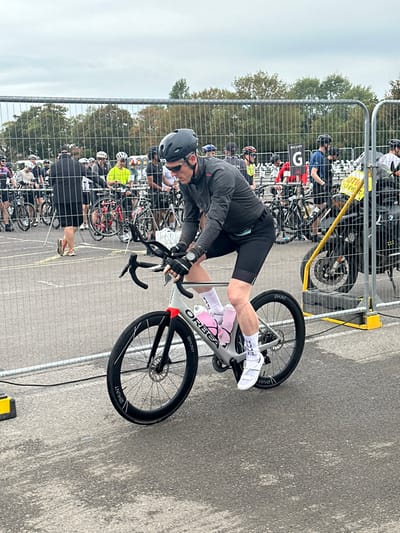 The image depicts an outdoor sporting event, likely a cycling race or triathlon, located in Weymouth, United Kingdom. The foreground features a male cyclist, appearing to be in his 40s or 50s, actively riding a modern grey Orbea aerodynamic road bike equipped with black Hunt wheels and disc brakes. He is dressed in a black cycling helmet, dark grey long-sleeved athletic top, black cycling shorts, white cycling socks with a visible "ICEC" inscription, white cycling shoes, and sunglasses, with a wrist-mounted device on his left arm. Two pink water bottles are secured in cages on his bike's frame.
Behind the cyclist, a temporary metal barrier separates the active course from a transition or staging area. This area is populated by numerous other individuals, presumed to be participants or event staff, standing near various bicycles. Visible elements in the background include bike racks, a white sign displaying "BOUVY" above a large "G", and a yellow sign partially reading "UTTO MOBILE EVENT". A section of a black motorcycle is also visible behind the yellow sign. The ground is a grey paved surface, and the sky is overcast. Trees are visible in the far background.