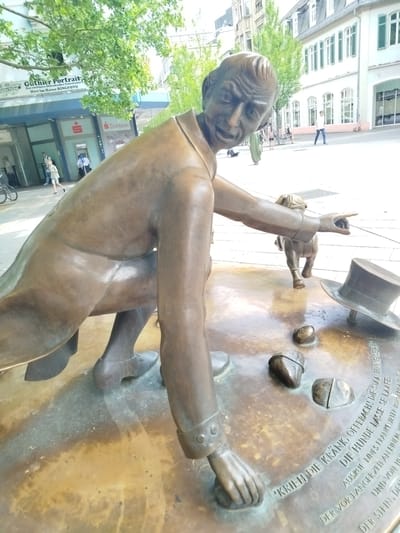 This media file captures a bronze statue of a man and a dog on a street in Offenbach am Main, Germany. The statue depicts a man in a suit, kneeling and pointing with his right hand, while a small dog, also bronze, walks away from him, seemingly being led by a leash. A top hat lies on its side on the ground near the statue. The base of the statue has an inscription in German.

In the background, a street scene unfolds with buildings and pedestrians. One building has a sign that reads "Guthier Portrait Das exclusive Offenbacher Fotostudio Hier im Hause RINGFOTO". Another building appears to be a "Sparkasse" (a German savings bank). People are walking in the background, and trees with green leaves suggest a pleasant time of day, likely daytime, with good weather. The overall mood appears to be a public space, possibly a square or pedestrian area.

The inscription on the base of the statue includes the phrases: "'KRIEH DIE KRANK, OFFEBACH! DIE STAA FINNE SEAL", "DER VOR LANGER ZEIT AN EINEM", "DIE HUNDE LASSE SE LAAFE.", "AUSRUF FINES FRANKFURTER BÜRGERMALINES", and "UND VON IH". The visible text and the "Sparkasse" sign provide clear location cues, identifying the setting as Offenbach am Main, Germany. The statue itself seems to be a public art installation.