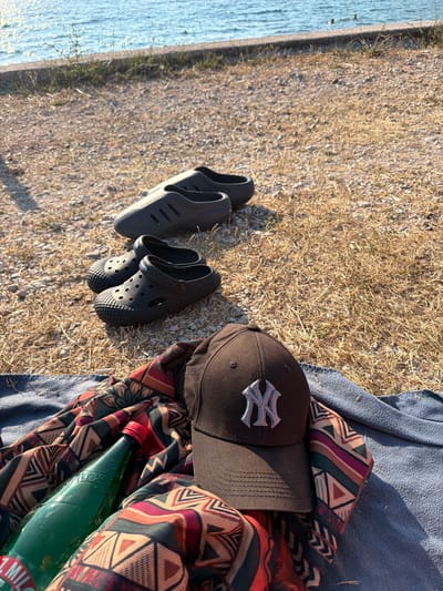 At the water's edge in Buljarica, Montenegro, a pair of black Croc-style clogs rests on dry, gravelly ground, positioned in front of another pair of black slip-on shoes. A brown New York Yankees baseball cap, emblazoned with a silver "NY" logo, is laid out on a blue towel. Adjacent to the cap, a green glass bottle with a red cap, partially obscured by a colorful patterned cloth, sits on the towel. The water, shimmering under sunlight, is visible in the background, with a low stone wall separating the dry ground from the sea. The overall scene suggests a relaxed day at the beach.