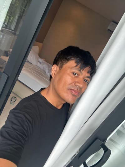 An Asian man in a black long-sleeve shirt poses indoors. He's positioned next to a window or sliding glass door with white curtains. The room's interior, including a bed with white linens, is partially visible behind him. The angle of the shot is slightly tilted. The frame of the window or door is black and modern.