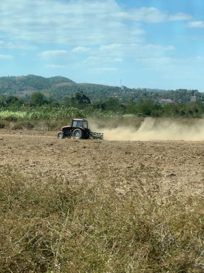 A red tractor tills a field, kicking up a cloud of dust. The field is brown and stretches into the distance. Lush green vegetation and a distant hill line the edge of the field. A blue sky with scattered clouds is visible in the background. The image is taken at an angle, making the scene appear slightly tilted.