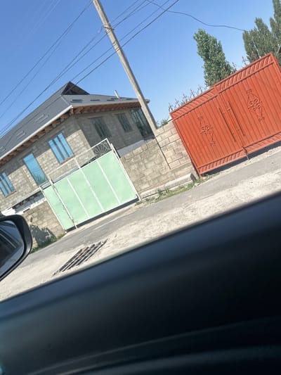 The image depicts a street scene in the city of Ынтымак, Kyrgyzstan, under a clear blue sky. On the left, a partially constructed building constructed from grey concrete blocks is visible. It has a dark grey, pitched roof with visible wooden beams. Several tall, rectangular windows with blue-tinted glass are present on this building. In front of the building is a large, light green metal gate, divided into four sections, with a white decorative element at the top.

To the right of the building, a tall, orange metal gate with decorative scrollwork is prominent. Behind the orange gate, several tall, slender trees with dense green foliage are visible. A concrete utility pole with multiple overhead wires runs diagonally across the upper portion of the image. The foreground of the image shows the interior of a vehicle, including a black car door and side mirror, and a section of the road with a metal grate. The scene appears to be captured from a moving vehicle.