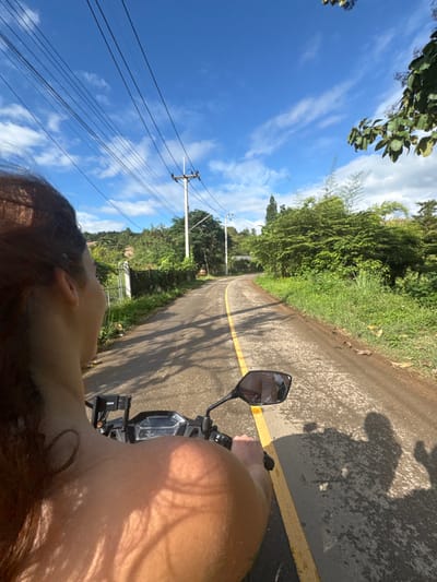 A first-person perspective image captures a scene along a winding paved road in Ban Mae Na Chalong, Thailand, under a clear blue sky with scattered white clouds. The foreground shows the back of a person's head and right shoulder, with brown hair appearing windblown. The person's skin shows a tan or sunburn pattern. Immediately in front of the person are the handlebars, dashboard, and right rearview mirror of a black scooter or motorcycle. A phone mount is attached to the handlebars, and a digital display on the dashboard shows "32."

The asphalt road features a solid yellow center line. Shadows from overhead foliage, and possibly the motorcycle and rider, are cast onto the road surface. Both sides of the road are bordered by dense green vegetation, including tall trees, bushes, and grass. Utility poles with multiple power lines run parallel to the road on the left. The road curves into the distance, continuing through a lush, somewhat hilly landscape.