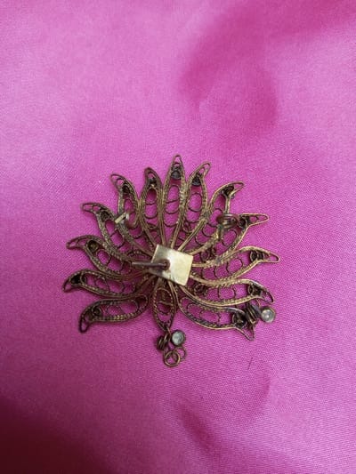 The image displays a single, intricate, metallic ornamental object, positioned on a magenta-colored fabric surface. The object, viewed from its reverse side, is circular in overall form, featuring approximately fifteen radiating, elongated lobes crafted from open-work filigree metal. The metal exhibits an aged, brass-like or bronze coloration with visible tarnishing.
At the center of the object's reverse, a flat, square, brass-toned metal plate is affixed, from which a straight, metallic pin extends diagonally towards the upper-left. Distributed across the reverse of the filigree lobes are multiple small, circular cup-like settings; some of these contain small, clear or white, circular components, while others appear empty. Towards the lower-right edge of the object, two small, interconnected metallic loops are visible, with one containing a similar small, white, circular component.
The background consists of a solid-colored fabric, which appears to be a magenta or deep pink material, possibly satin or silk, showing slight creases and a subtle texture. The scene is illuminated by diffuse, ambient light, producing no harsh shadows. No individuals, discernible actions, or interactions are depicted. The object is reportedly from Qarabucaq qəsəbə, Azerbaijan, though no visual context for this location is present within the frame.