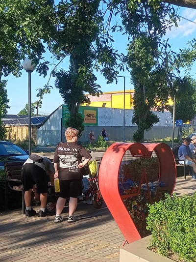 The image depicts an outdoor scene in Bilhorod-Dnistrovskyi, Ukraine, likely on a sunny day, with lush green trees providing shade. In the foreground, two individuals are visible. One, from the back, is a woman wearing a black t-shirt with "BROOKLYN" printed on it in large white letters and black shorts. She is holding a yellow bottle in her right hand. Beside her, a person is bent over, seemingly attending to something near a bench. A portion of a black car is visible to the left. Further into the scene, a distinctive bright red, heart-shaped trash receptacle is prominent, filled with various items. In the background, there are buildings, one with solar panels on its roof, and a large yellow structure. A street sign and a pedestrian crossing sign are also present. Several other individuals are seated on benches in the distance, observing the surroundings. The ground is paved with patterned tiles.