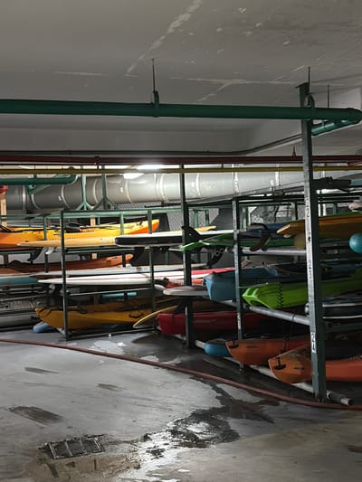 The image shows a storage area for kayaks and surfboards in a building. There is a metal rack system holding the watercraft, which are of various colors and sizes. A large, segmented ventilation duct runs parallel to the rack. The location appears to be an indoor storage facility. The presence of water on the floor and a floor drain suggests this area may be prone to moisture or drainage issues. There is no indication of people or interaction in the image. No evidence directly connects the scene to Key Biscayne, United States of America.