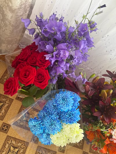 The image displays four bouquets of flowers on a patterned rug. The bouquet in the top left contains red roses, while the bouquet directly above it is composed of purple bellflowers. To the right of the bellflowers, dark red lilies are visible, and below them are orange lilies and other assorted flowers. In the bottom left, a bouquet features blue chrysanthemums and white chrysanthemums. The scene is indoors, with a sheer curtain in the background. There are no people or animals present in the image, and no specific actions or interactions are depicted. The environment appears to be a room, possibly a home, given the rug and curtain. The location of Елимбетово, Russia is not visually ascertainable from the provided image.