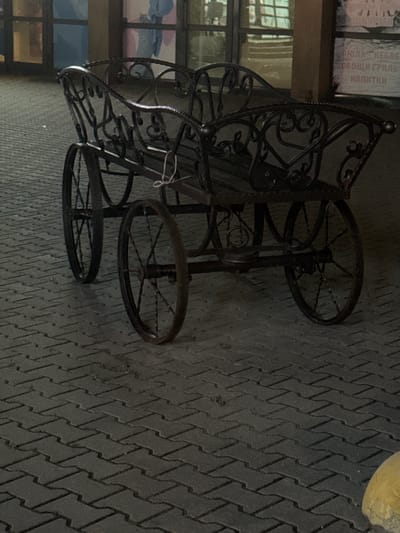 An ornate, dark metal, decorative carriage-like object with two large wheels in the back and two smaller wheels in the front rests on a gray, interlocking brick paved ground. The carriage has decorative scrollwork on its sides and backrest. A thin white string is tied to the left side of the carriage. In the background, there are large windows, some of which show reflections and muted colors. To the right of the carriage, a vertical brown pillar is visible, and a poster with text, possibly in Cyrillic, is affixed to a surface behind it. The lighting suggests it is either evening or indoors with artificial lighting. The environment appears to be an urban or public space.