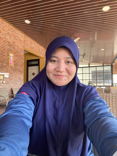 In Keraton, Indonesia, a young woman wearing a dark blue hijab and a matching long-sleeved shirt is captured in a close-up selfie. She is seated on a wicker chair on what appears to be a patio or veranda, with a brick wall and a wooden-paneled ceiling visible in the background. The setting suggests an outdoor or semi-outdoor environment, possibly a cafe or a home's outdoor living area, given the presence of a ceiling fan and glass-paneled doors. The woman is looking directly at the camera with a slight smile, her face well-lit by natural light. Her attire is modest, and her expression is pleasant. The background elements, such as the brick wall with a framed picture and an electrical outlet, provide context to the location. The focus of the image is on the woman, with the environment serving as a backdrop.