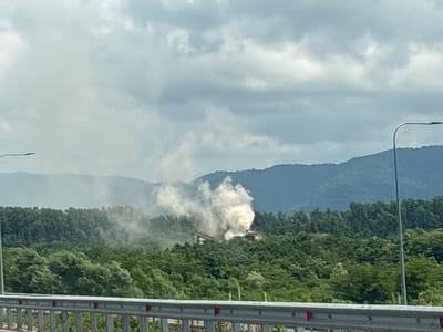 The image depicts a landscape view from a bridge or overpass, with a metal railing in the foreground. Beyond the railing, a dense forest covers rolling hills, leading to larger, mist-shrouded mountains in the background under a cloudy sky. In the middle ground, amidst the trees, a industrial facility is visible, emitting a large plume of white smoke. The facility appears to be a structure with elevated conveyor belts or pipes and some yellow machinery. The overall scene suggests an industrial operation within a natural environment, with the smoke indicating ongoing activity. The specific location is identified as the city of Lanchkhuti, Georgia.