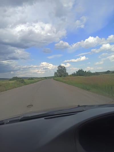 The image captures a scenic view from a vehicle traveling on a dirt road, likely in Novoselivka, Ukraine. The perspective is from inside the car, with a portion of the car's interior visible on the right side of the frame. The scene is dominated by a vast, partly cloudy sky with a mix of bright blue and dramatic white and gray clouds. To the left of the road, a field of tall, dry grass extends towards a line of trees and more distant vegetation. The dirt road itself is a light brown color and appears to be well-maintained. The lighting suggests it is daytime, possibly late afternoon or early evening, given the soft light. There are no people visible in the image, and no text is present. The overall impression is one of a peaceful, rural landscape on a day with dynamic weather.