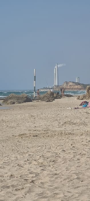 The image shows a sandy beach in the city of Mihmoret, Israel. The coordinates 32.409, 34.869 correspond to the location. The beach is mostly empty, with a few people in the distance. There are also a few small rocks scattered on the sand. In the background there is a tall chimney with smoke coming out of it. There is also a  building in the distance. The sky is blue and cloudless. The beach is located near a power plant. The power plant is visible in the distance, with two large chimneys and a structure nearby.  There is a black and white striped post standing upright in the sand. There are a few people standing in the sand near the waterline. The beach is located in a relatively isolated area and surrounded by sand dunes. There is a small amount of debris scattered along the shoreline.  A few individuals are walking along the beach, moving away from the waterline. There is also a group of people in the distance gathered around the power plant.  The image appears to be taken on a clear day.