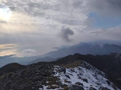 The image displays a wide-angle panoramic landscape captured from a high mountainous vantage point in the vicinity of Mali Mikulići, Montenegro. The immediate foreground features a rugged, rocky ridge with patches of white snow interspersed among dark grey stones and sparse, dry brown vegetation. Beyond this, successive layers of dark, undulating mountain ranges, appearing forested or heavily vegetated, recede into the distance, partially veiled by atmospheric haze and low-lying clouds.
On the left side of the frame, the Adriatic Sea stretches towards the horizon, its surface reflecting diffused sunlight that penetrates breaks in an otherwise overcast sky. Along the coastline, a dense urban area, characterized by numerous buildings and visible harbor infrastructure, is situated at the base of the mountains. The sky overhead is largely obscured by a dense layer of grey and white stratocumulus clouds. A brighter, yellowish-white glow in the upper-left indicates the sun's position behind the clouds, while a distinct, dark grey, vertically elongated cloud formation is prominent slightly left of the image's center, mid-sky. A small segment of blue sky is visible in the upper-right quadrant. No individuals, animals, or specific man-made objects other than the distant urban landscape are present. No actions or interactions are depicted within the scene.