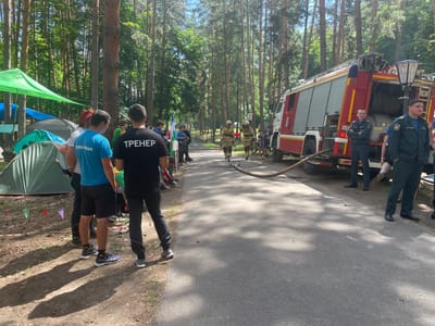 The image depicts a scene in Nikolayevka, Russia, with a focus on a fire and rescue training or competition event. The setting is a wooded area with a paved road. A fire truck, red and white, is prominently parked on the right side of the road. Several individuals are present, some in what appear to be firefighter uniforms, and others in casual athletic wear. Two people in full firefighting gear are walking on the road, and one is holding a hose connected to the fire truck. In the background, tents are set up, suggesting a temporary camp or event setup. The overall atmosphere appears to be active and organized, likely related to a local event or competition.