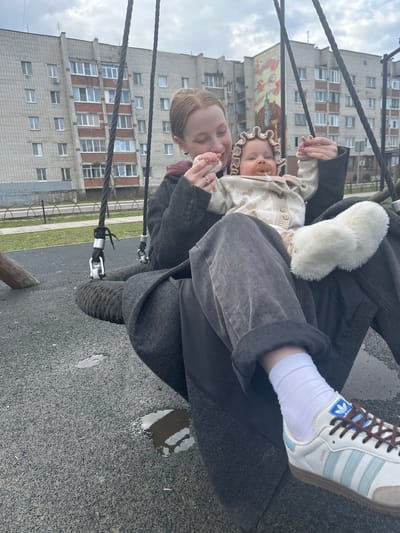 A medium shot shows a light-skinned woman on a playground swing, holding a baby. The woman wears grey jeans, a grey coat, white socks, and white Adidas Samba sneakers with brown laces and blue stripes. The baby, wearing a tan bonnet and patterned beige cardigan, is held by the woman who is holding the baby's hand with her left hand. The building in the background is a grey apartment building with a mural painted on the left side. The playground has a grey rubber surface with puddles. The location is potentially Pokrov, Russia.