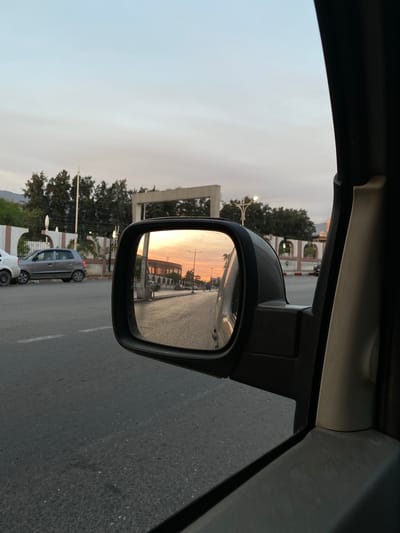 This image, taken from inside a vehicle, captures a street scene in Blida, Algeria, during what appears to be sunset or sunrise. The view is framed by the car's interior, with a black side mirror prominently displayed. The mirror reflects a road lined with buildings and trees, bathed in the warm, orange glow of the setting or rising sun.
In the foreground, the asphalt road shows white lane markings. To the left of the mirror, a silver car is parked, and a white car is partially visible behind it. Beyond the parked cars, a low, white wall with decorative arches runs along the street, backed by a line of trees. In the reflection of the mirror, the road continues into the distance, with more buildings and streetlights visible. The sky in the reflection is a soft blend of orange and pink, indicative of twilight.
The overall environment suggests a city street, possibly a residential or commercial area, experiencing a transitional time of day. The image has a slightly grainy texture, common in photographs taken with mobile devices or in low light conditions.
