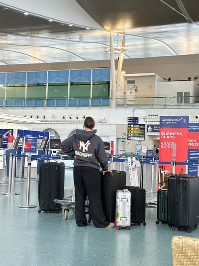 In a bright, modern airport terminal, a person stands facing away from the camera, looking towards a check-in counter. They are wearing a dark grey hooded sweatshirt with a New York Yankees logo and the text "MADHAPPY" printed on the back, along with black pants and light-colored shoes. The person is surrounded by several pieces of luggage: two large black suitcases on their left, a smaller silver suitcase with colorful stickers, a medium-sized black suitcase, and a dark green suitcase on their right. A luggage cart is also present near the person.

In the background, rows of check-in counters are visible, some with Aeroflot branding and signage indicating "BUSINESS CLASS" and "SKY PRIORITY." A large screen displays airport information, and automated check-in kiosks are also present. The airport architecture features a high, vaulted ceiling with metal beams and lighting fixtures. The floor is a light blue speckled surface. The scene appears to be within the public area of an airport, possibly the departure hall. The location is identified as Tambon Mai Khao, Thailand, indicating this is likely an international airport terminal within that region.