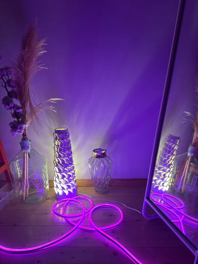 In a room with purple lighting, a wooden floor is visible. On the floor, a bright pink neon light strip is arranged in loops. To the left, a large clear glass vase contains dried pampas grass and what appears to be cotton branches, illuminated from within. Next to it is a tall, textured lamp with a pattern that resembles scales, emitting a yellow and purple light. Further to the right, a smaller, faceted glass vase is placed on the floor. A large mirror on the right side of the frame reflects the scene, including the objects and the purple light. The overall ambiance is dimly lit and atmospheric, with the neon light providing a strong visual element. The location is specified as Буран Снт, Russia.