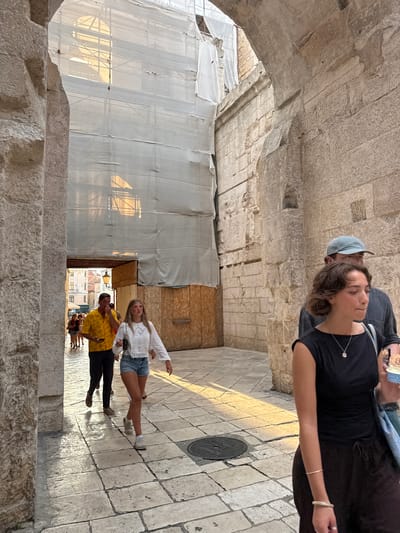 This image depicts a street scene within the Diocletian's Palace complex in Split, Croatia. The foreground features a woman in a black sleeveless top and dark trousers, wearing a silver necklace and bracelets. She is walking to the right, with a blue bag slung over her shoulder and holding a light blue cup. Behind her, partially obscured, is a man wearing a blue baseball cap and a dark shirt, also walking to the right.
In the mid-ground, a young woman with blonde hair is walking to the left, wearing a white lace top and denim shorts. To her left, a man in a bright yellow patterned shirt and dark trousers is also walking left, looking down at his phone. Further in the background, other pedestrians are visible walking down the narrow street.
The environment is characterized by ancient stone architecture, with large stone blocks forming the walls and arches. The street is paved with stone tiles. Overhead, scaffolding and white sheeting are present, suggesting ongoing construction or restoration work. Sunlight casts strong shadows and highlights on the stone surfaces, indicating it is either early morning or late afternoon. The scene captures a moment of daily life within the historic palace grounds.