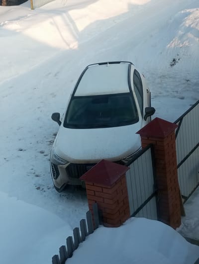A white sport utility vehicle (SUV) is parked on a snow-covered surface in город Яромаска, Russia, observed from a high-angle perspective. The SUV's front grille and lower bumper are soiled, while its white body, roof rails, and sunroof appear cleaner. To the right of the vehicle, a fence line is present, featuring two brick pillars topped with red caps, partially obscuring a corrugated metal gate. A lower, dark-colored picket-style fence is visible in front of the pillars, partially covered by a substantial accumulation of snow. The ground across the entire scene is uniformly blanketed in snow, with minor disturbances or tracks discernible near the vehicle. Bright daylight illuminates the environment, casting shadows on the snow-laden background terrain. No individuals or animals are present in the scene.