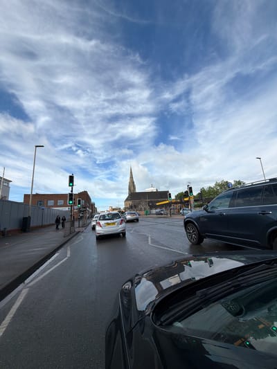 This image depicts a bustling urban street scene in Leicester, United Kingdom, during daylight hours. The road appears wet, suggesting recent rain. The perspective is from the roadside, with the glossy dark body of a vehicle occupying the right foreground, reflecting the street and surrounding structures.
Along the street, several cars are visible, including a white vehicle with the UK license plate "FN62 XWR" in the mid-ground. Multiple traffic lights are strategically placed, all illuminated green, indicating an open flow of traffic. Pedestrians are scattered along the sidewalk, going about their daily activities.
Architecturally, the scene features a prominent church spire rising in the distance, a common landmark in British towns and cities. Other buildings lining the street vary in style, with one building displaying the partial text "STUDIOS." The sky overhead is a clear blue, adorned with wispy white clouds, creating a bright atmosphere. Street furniture, such as lampposts and a blue circular sign, are also part of the urban landscape. The overall impression is one of an active, everyday city environment.