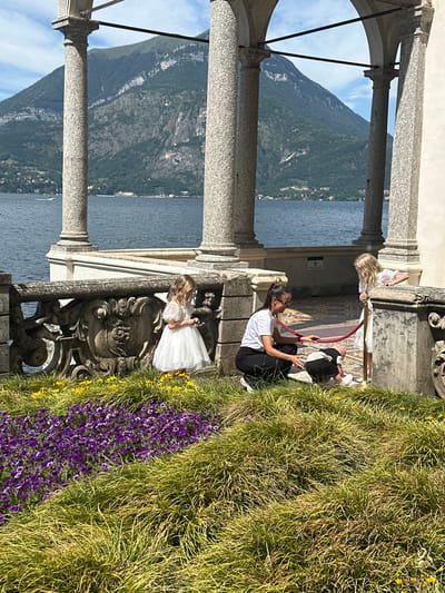 In Varenna, Italy, a woman in black pants and a white t-shirt kneels on the ground, interacting with a child in a black and white outfit. Nearby, a young girl in a white dress stands to the right, looking at the child and woman. They are positioned on a stone patio with a ornate stone railing overlooking a body of water and mountains. A vibrant flowerbed of purple and yellow flowers, with tall green grass, is in the foreground. The scene is framed by large stone columns and arches.