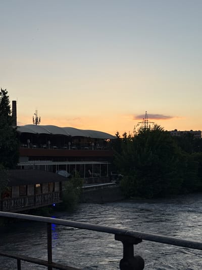 This vertical, eye-level, medium shot depicts a riverfront scene at dusk, likely in Astrakhan, Russia. The sky is a gradient of pale blue transitioning to warm orange near the horizon. In the middle ground, a large, multi-story building with a distinctive arched, covered roof extends across the frame. This building appears to be a restaurant or entertainment venue, with tables and chairs visible on its upper level. A tall, brick smokestack stands to the left of this structure. To the right of the main building, a dense cluster of dark green trees is prominent. A lattice-like electricity pylon stands amidst the trees, silhouetted against the colorful sky. In the foreground, a dark metal railing runs horizontally across the lower portion of the image, obscuring a portion of the river. The river itself is a turbulent, dark gray, with visible ripples and currents. A small, dark wooden structure with windows and a railing, possibly a covered walkway or a small building, juts out over the river on the left side, with a faint blue light visible beneath it. Buildings and trees are also visible on the far bank of the river, appearing as dark shapes against the twilight sky. No people are clearly identifiable in the image. The overall impression is one of a relaxed, evening ambiance, with the natural elements of the river and sky contrasting with the man-made structures of the city.