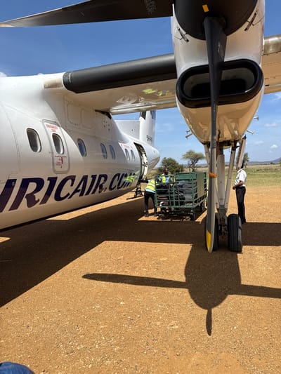 A partial view of an airplane on a brown surface shows the propeller, nose, and side of the aircraft. The airplane is white with "AFRIC AIR.COM" printed on its side in dark blue. A luggage cart is adjacent to the plane, with ground crew nearby. A person in a pilot's uniform stands near the plane's wheel, while the shadow of the propeller extends across the ground. The sky is visible in the background.
