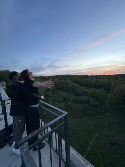 The image depicts two men sharing an intimate moment on a high viewpoint, gazing out over a forested landscape under a twilight sky.
In the foreground, one man with dark skin, short dark hair, wearing a dark top and olive green pants, stands with his back partially to the camera, looking upwards. His left arm is down, and he wears white sneakers. Leaning against him, with his head resting on the first man's shoulder, is a second man with lighter skin, a beard, and glasses. He wears a black t-shirt and black pants. His arms are outstretched wide, palms facing upwards towards the sky, with a lit cigarette held between the fingers of his right hand. A ring is visible on his left ring finger. Both men appear to be absorbed in the vast view and the sky above. A black bag is visible near the first man's feet.
They are positioned behind a dark, somewhat weathered metal railing, possibly part of a bridge, viaduct, or observation deck, which frames the lower portion of the scene. Below the viewpoint, a dense, dark green forest stretches across rolling terrain, fading into the distance. Beyond the tree line, on a sloping expanse of grass, faint, winding paths are discernible.
The sky, dominating the upper part of the image, displays the beautiful hues of dusk or dawn. It transitions from a soft blue on the left to gentle pinks, oranges, and pale yellows towards the horizon, indicating the sun has either just set or is about to rise. The overall atmosphere conveys a sense of peace, freedom, and shared wonder. No visible text is present in the image.