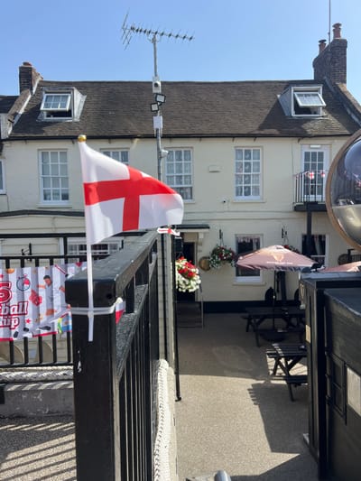 The image shows the exterior of a building, possibly a pub or restaurant, decorated with English flags and banners. The building is white with multiple windows, and a dark-tiled roof. An open window on the upper floor reveals a glimpse of the interior. There are flower arrangements on balconies. In the foreground, an English flag is prominently displayed, attached to a black railing. Below the railing, several colorful banners are visible, with some text partially obscured. One banner appears to have text related to "Ball" and possibly "Championship". The scene suggests a celebration or a festive occasion, possibly related to a sporting event or national holiday, given the presence of the English flag and celebratory banners. The sunlight indicates it's daytime. The setting appears to be outdoors, with a clear blue sky visible. The overall mood is one of patriotism and festivity.