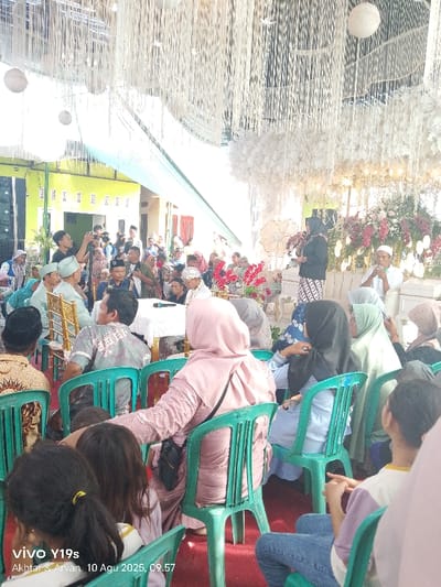 The image captures a formal gathering or ceremony in Jagapura Kulon, Indonesia, on August 10, 2025, at 09:57. The setting is a semi-outdoor event space, indicated by a temporary canopy structure adorned with white streamers, white spherical ornaments, and light blue fabric drapes suspended from the ceiling.
At the far right, a raised stage or focal area is decorated with extensive floral arrangements featuring white and reddish-pink flowers. A woman dressed in a black top, a patterned skirt, and a black hijab stands on this stage, actively speaking into a microphone. To her immediate right, a man in a white shirt and a white peci (skullcap) is seated.
Multiple attendees are seated in rows of green plastic chairs, primarily facing the stage. In the foreground, a woman in a light pink hijab and a textured pink dress is seated with her back to the viewer, a black shoulder bag resting on her chair. Other seated individuals, predominantly women wearing various colored hijabs (black, light blue, green, grey, brown) and men in batik-patterned shirts, are visible. A white-covered table with a microphone rests among the seated guests in the mid-ground.
In the left background, a lime-green building with windows is visible. Several individuals stand in this area, with one person holding a camera or smartphone, seemingly recording the event, and another holding a microphone. The ground beneath the seated area is covered by a red carpet. The scene suggests a community event or celebration, likely a wedding or similar formal occasion.