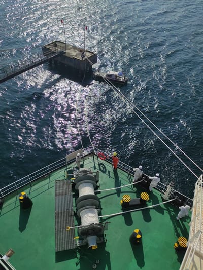 This high-angle, top-down image captures a marine operation taking place off Kunak, Malaysia, under bright daylight with strong sun reflections on the water. The scene features the green deck of a large vessel, bustling with activity, alongside two smaller marine craft.

On the main ship's deck, several individuals are visible. Three people wear full white protective suits, positioned near black bollards with yellow safety stripes. Another person, dressed in an orange long-sleeved shirt and a yellow hard hat, is seen closer to the edge of the vessel, seemingly involved in overseeing the operation. Dominating the foreground of the deck are large winches or capstans with heavy white ropes neatly coiled around them. Metal grates and additional black bollards are also present on the green deck.

Connected to the main vessel by multiple ropes are two smaller craft. To the left, a square, dark-colored floating platform or barge is visible, featuring a ladder extending into the water and two poles with red tips. Closer to the main ship, a white "PILOT" boat is positioned alongside, its name clearly visible on its side. A person can be seen on board the pilot boat.

The water surface is rippled and heavily reflects the sunlight, indicating a clear, sunny day. Sharp shadows cast by the ship's structures and equipment further confirm the bright conditions. Visible text on the main ship's deck includes "SWL 7.2S" and "SWL 7.2 TON 03E" near some bollards, and "FD" inscribed on a railing. The overall activity suggests a coordinated effort involving personnel transfer or specialized marine work.