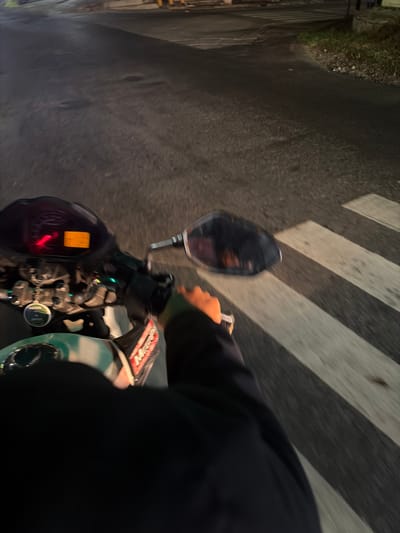 An over-the-shoulder, first-person perspective shot depicts a motorcycle rider at night, illuminated by streetlights and the vehicle's dashboard. The rider, clad in dark clothing, is captured from the chest down, with a gloved hand visible on the handlebars. The motorcycle's instrument panel, featuring a red-lit tachometer and an orange digital display, is to the left. A side-view mirror, reflecting the night sky and faint lights, is positioned to the right. The motorcycle is in motion, indicated by the blurred road surface and the motion blur on the pedestrian crossing ahead, which features stark white stripes. The background consists of a dark asphalt road with some irregularities and patches, and a patch of overgrown green grass to the right, bordering the road. The scene is set in a dimly lit urban environment, possibly on a street in Boja, Indonesia, at night.