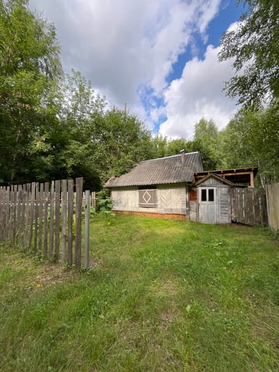 A weathered, wooden house sits in a grassy yard surrounded by trees, under a cloudy sky with patches of blue. A rustic wooden fence encloses part of the yard. The house has a grey, corrugated roof and light-colored wooden siding with a decorative pattern. There is a small attached structure with a wooden door and a covered open area. A single window with diamond-shaped shutters is visible on the main house. The scene appears to be located in Nagornyi (Sitnikovskii S/S), Russia.