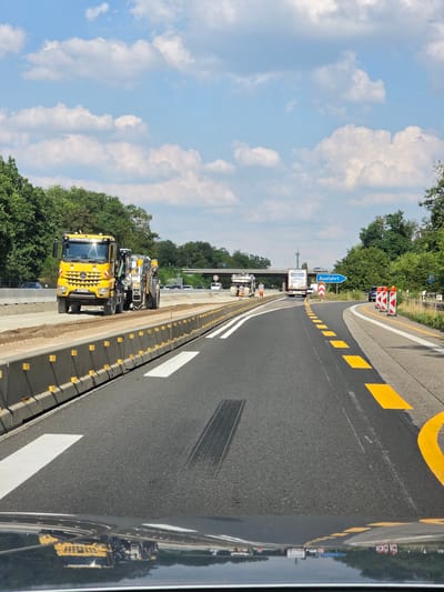 The image depicts a construction zone on a highway in Rüsselsheim am Main, Germany. A large yellow road milling machine, branded "LEONHARD WEISS," is actively working on the road surface. The machine appears to be removing material from the existing asphalt. In the background, other vehicles, including cars and trucks, are visible on the highway, some of which are navigating through the construction area. A blue directional sign indicating "Ausfahrt" (Exit) is present, suggesting an upcoming exit from the highway. The scene is characterized by temporary concrete barriers lining the construction area, road markings, and traffic cones, all typical elements of highway construction. The sky is blue with scattered white clouds, and lush green trees line the sides of the highway. The reflection on the dark surface in the foreground suggests the photo was taken from a vehicle.