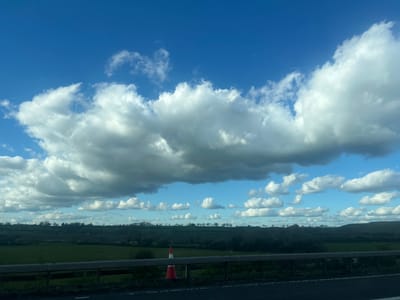 The image depicts a bright blue sky with thick, white cumulus clouds. In the background, a landscape is visible, including green fields, trees, and a dark stretch of pavement. A red and white striped object is visible next to the road. The image is upside down. This image likely originates from Royal Wootton Bassett, United Kingdom, based on the context provided.