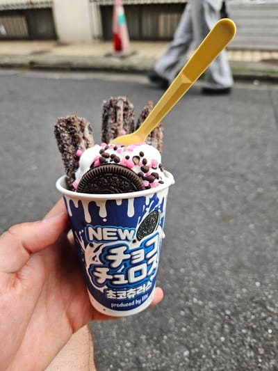 A person holds a cup of Korean ice cream and churros in their hand. The cup is labeled with Korean writing and the English word "NEW," and contains a dessert with whipped cream, chocolate sprinkles, and an Oreo cookie. Several churros stick out from the top of the cup. A yellow plastic spoon is inserted into the cup. The person is standing on a paved street. Blurred background elements suggest an urban environment, with a building and person visible. Based on context, this image was likely taken in Shinjuku, Japan.