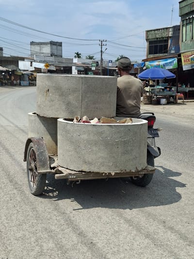 In Keudegeudong, Indonesia, a person wearing a beige shirt and a cap is riding a motorcycle that is pulling a trailer loaded with large concrete cylinders. The trailer appears to be homemade, constructed from metal and wood, with a single wheel visible on the left side. The concrete cylinders are stacked, with two full cylinders and one partially filled with what appear to be paper or fabric scraps. The motorcycle has a license plate that reads "92 KAN 08-24". The scene takes place on an asphalt road, with buildings and market stalls lining the street on the right side. Overhead wires crisscross the sky, and the weather is clear with a bright sun casting shadows. The overall impression is one of everyday commerce and transportation in a bustling Indonesian town.