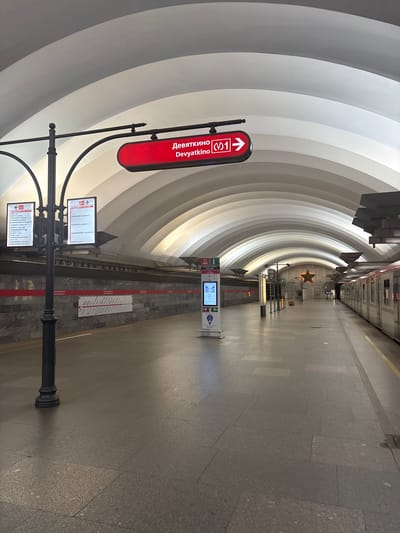 The image depicts the interior of a subway station, specifically the Devyatkino station in Kokkorevo, Russia. The station features a long, curved, vaulted ceiling that is white and illuminated by soft lighting. A train is visible on the right side of the frame, stopped at the platform. The platform itself is made of stone tiles and is empty of people.
Prominently displayed on the left side of the platform is a directional sign with red and white lettering indicating "Devyatkino" and the Metro line number "1" with an arrow. Above this sign, on a lamppost, are informational boards displaying subway maps and station names. Further down the platform, a digital information kiosk is present.
The walls of the station are clad in stone, with a red stripe running horizontally along them. In the distance, a large golden star is mounted on the wall, a common architectural element in some Russian metro stations. The overall impression is one of a clean, modern, and well-lit public transportation hub, devoid of any human activity at the time the photograph was taken.