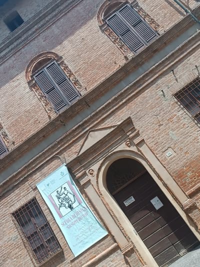 The image shows the exterior of a brick building, likely in Casalmaggiore, Italy, featuring architectural details such as arched windows with wooden shutters and decorative brickwork. A banner is hung on the left side of the building, displaying text that reads "SCUOLA DI DIS" and "GIUSEPPE BOTTO". The building has a large wooden door with a small plaque on it. A number "29" is visible on the brick wall near the door. The perspective is from a low angle, looking up at the building, with the scene tilted.