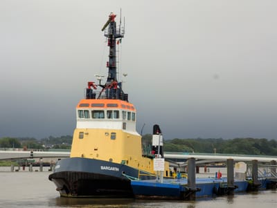 This media file shows a tugboat named "BARGARTH" docked at a pier. The tugboat is a prominent subject, with its yellow and orange superstructure and dark blue hull clearly visible. It is attached to a blue barge or pontoon.

The scene is set on a body of water, likely a river or harbor, given the presence of a bridge in the background and lush green trees and buildings lining the far bank. The weather appears overcast and potentially rainy, with a dark, stormy-looking sky dominating the upper portion of the frame.

Notable details include the various pieces of equipment on the tugboat's mast, including radar, lights, and a flag pole with an Irish flag. There are also mooring ropes connecting the tugboat to the pier. A "NOTICE" sign is visible on the side of the barge, with text that reads: "PRIVATE BERTH / NO PUBLIC MOORING / NO ACCESS TO SHORE / Port of Waterford Company".

The time of day is not explicitly stated, but the overcast conditions suggest it could be daytime. The overall mood is somewhat somber due to the weather, but the presence of the docked vessel and the bridge indicates ongoing activity or infrastructure in the area. No people are visible in the image.