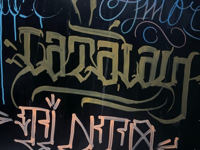 A close-up, horizontal shot captures a black surface adorned with graffiti. The graffiti features stylized lettering in yellow and orange, with some blue and pink accents in the upper portion of the image. The yellow lettering is large and appears to be the primary focus, characterized by thick, flowing strokes. Beneath this, the orange graffiti is also stylized but in a more angular, blocky fashion. The background is uniformly black, suggesting it might be a wall or a blackboard. The image's composition is tightly cropped, focusing solely on the graffiti art, and does not provide any contextual information about its specific location within Santa Marta, Colombia, or any identifiable people or actions.
